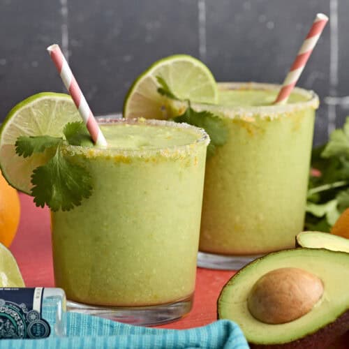 Side view of two glasses of avocado margarita, garnished with lime slices and fresh cilantro.