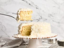 A piece of coconut cake being taken from a cake stand.