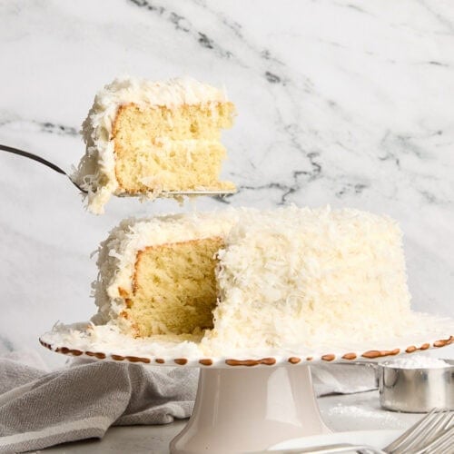 A piece of coconut cake being taken from a cake stand.