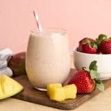A glass of homemade strawberry mango smoothie with a straw, on a wooden cutting board with strawberries and mango chunks.