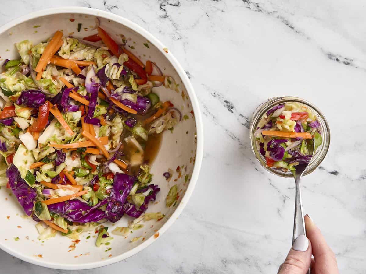 Prepped and massaged vegetables in a mixing bowl, with a spoon portioning some into a glass mason jar for fermentation.