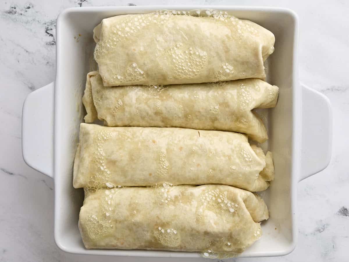 Folded chicken chimichangas in a baking dish, topped with cooking oil spray.