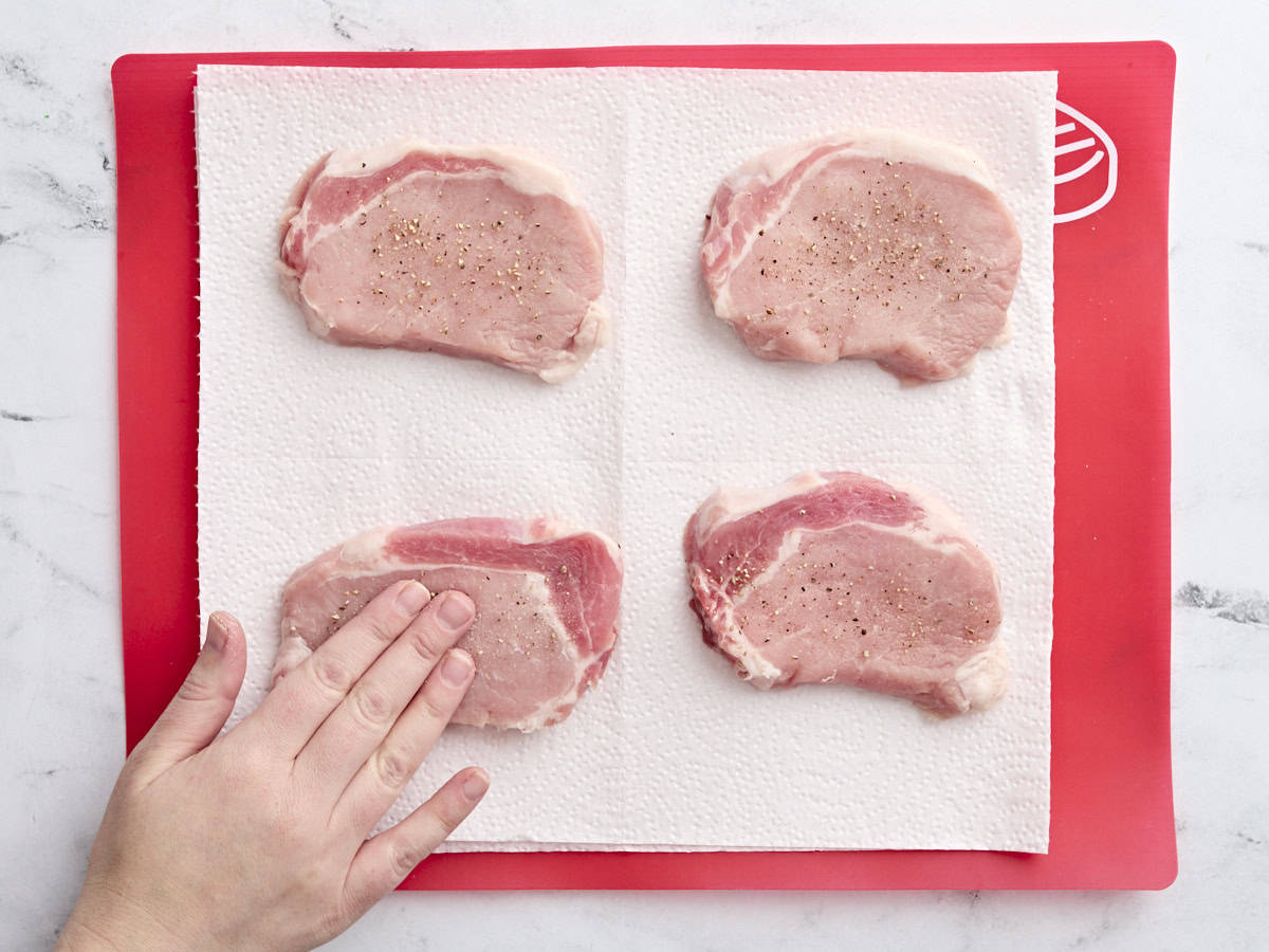 A hand rubbing salt and pepper onto a raw pork chop.