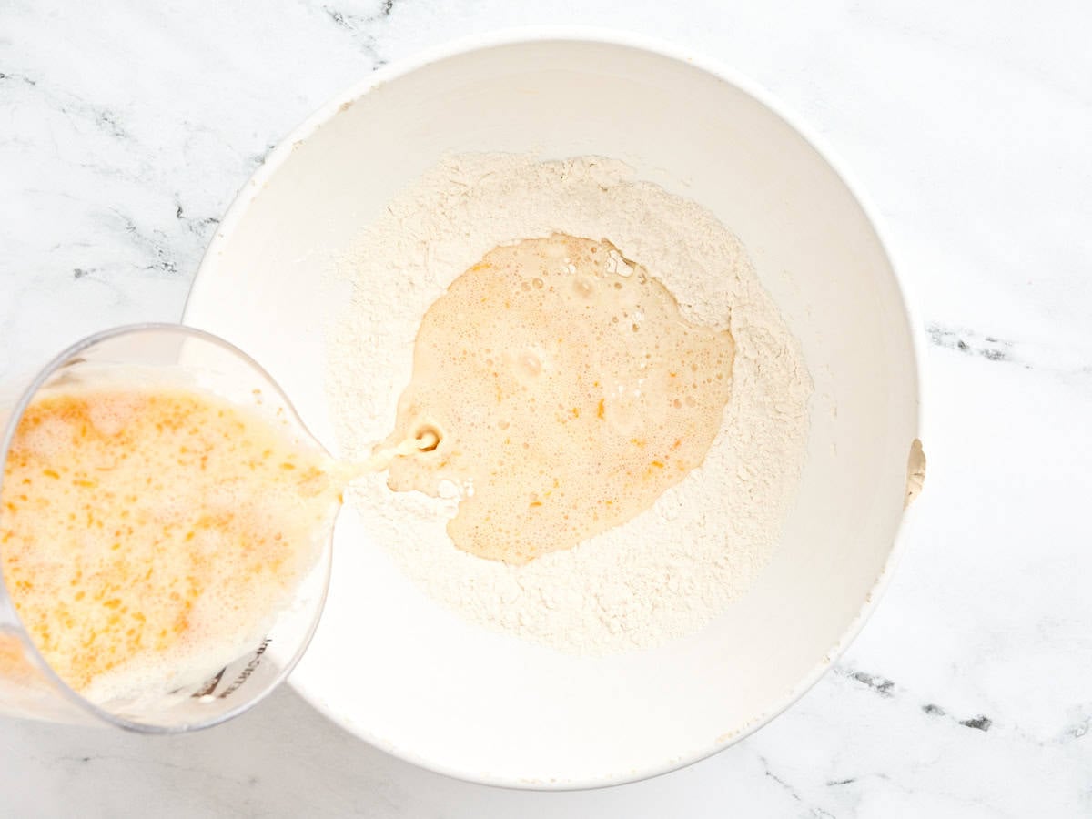 A yeast mixture being poured into the bowl of dry ingredients to make a dough for orange rolls.