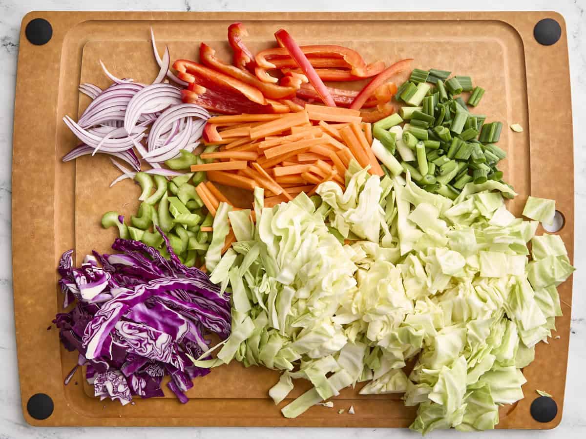Sliced green cabbage, sliced red cabbage, sliced green onions, chopped carrots, red bell pepper and red onion on a wooden cutting board.