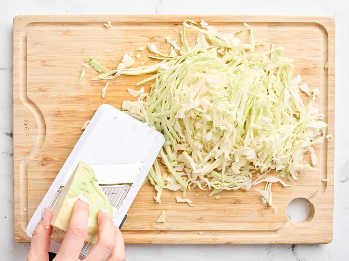 Hands shredding white cabbage with a mandolin slicer.