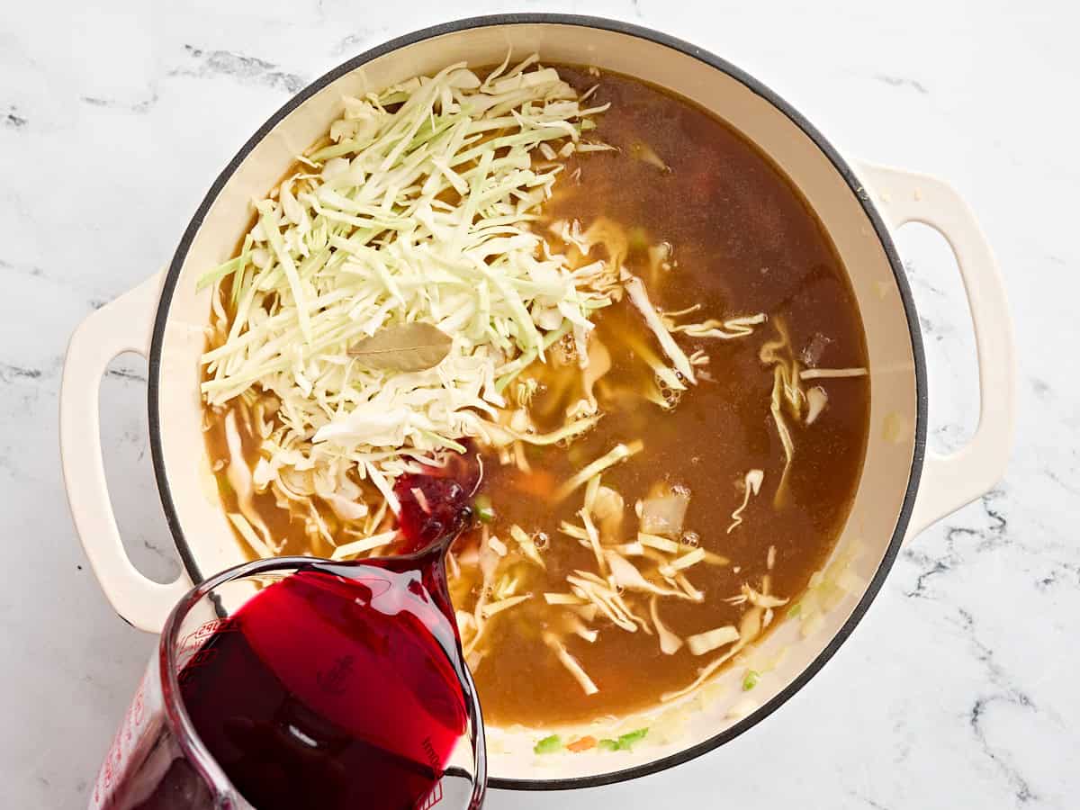 Broth, shredded cabbage and beet juice being added to sautéed mirepoix in a sauce pan.