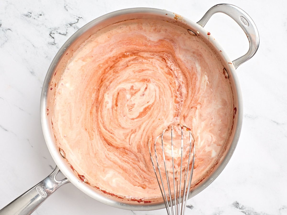 Heavy cream being whisked into tomato paste in a skillet.