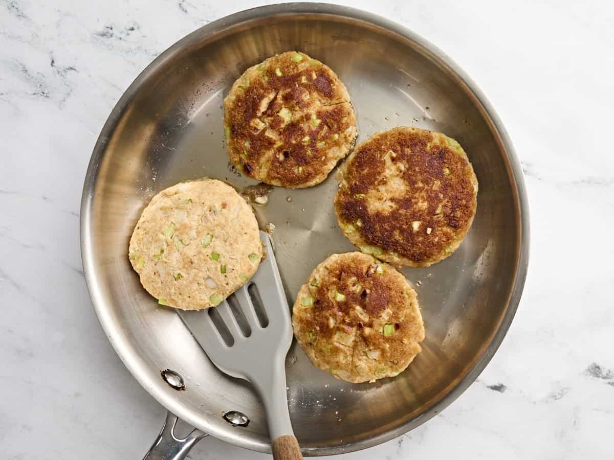Homemade crab cakes cooking in a skillet, with a spatula about to flip one over.