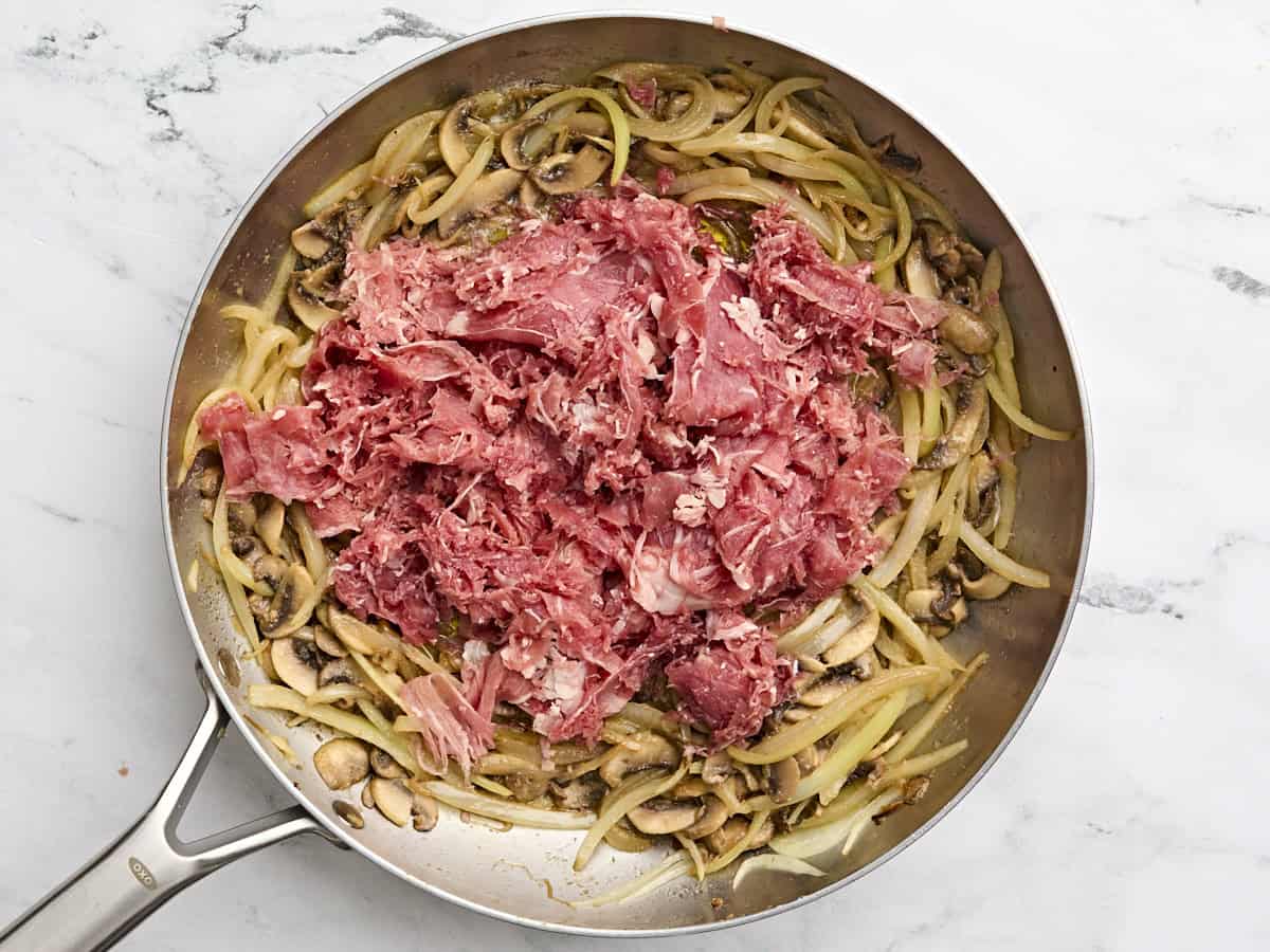 Shaved beef steak added to sauteed onions and mushrooms in a skillet.