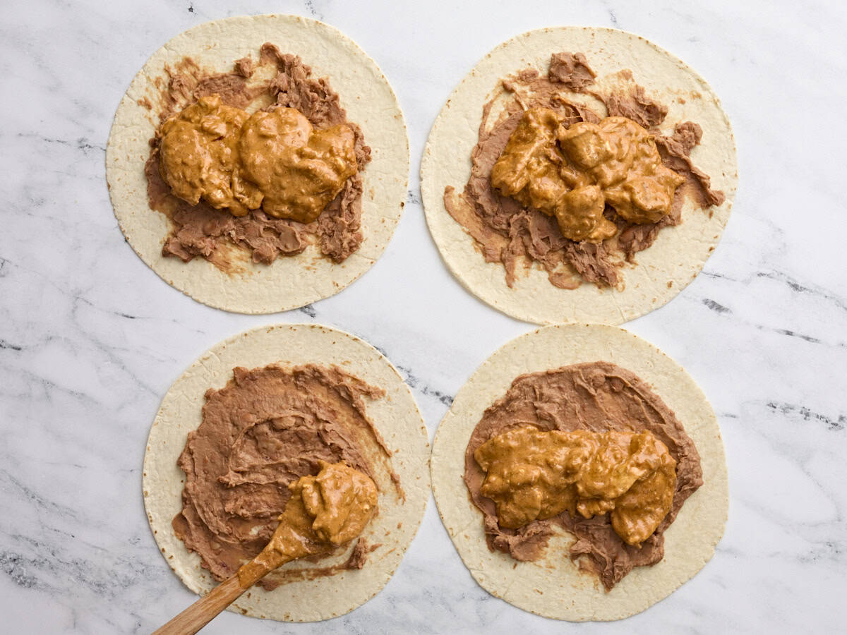 Four flour tortillas lay flat on a work surface, with refried beans spread on top and a spoon adding chicken chimichanga filling on top.