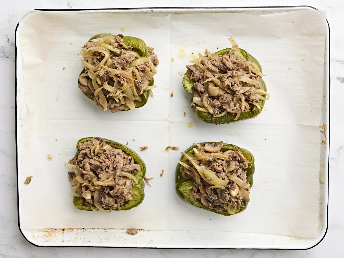 Green bell pepper halves filled with cooked beef steak, onions, and mushrooms on a baking sheet.