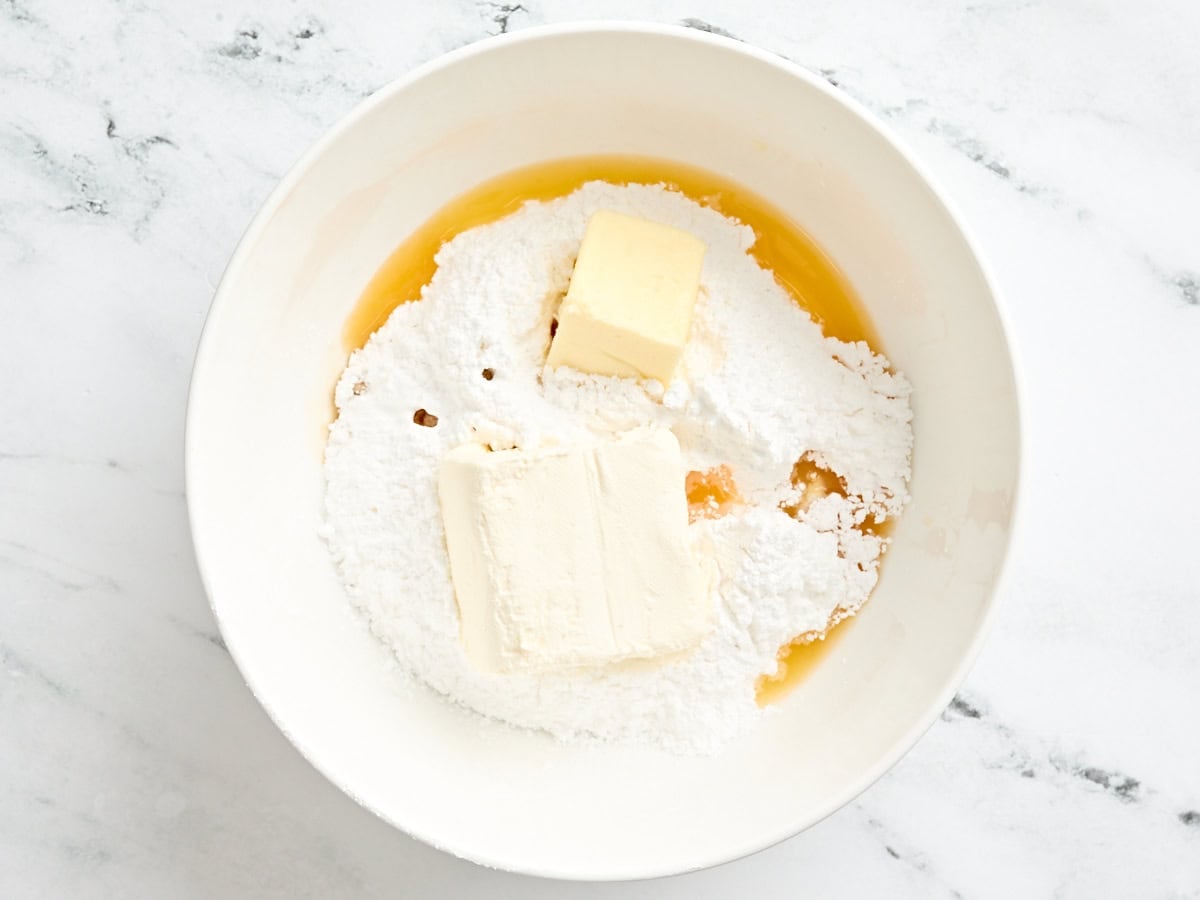 Powdered sugar, butter, orange juice, and cream cheese in a mixing bowl.
