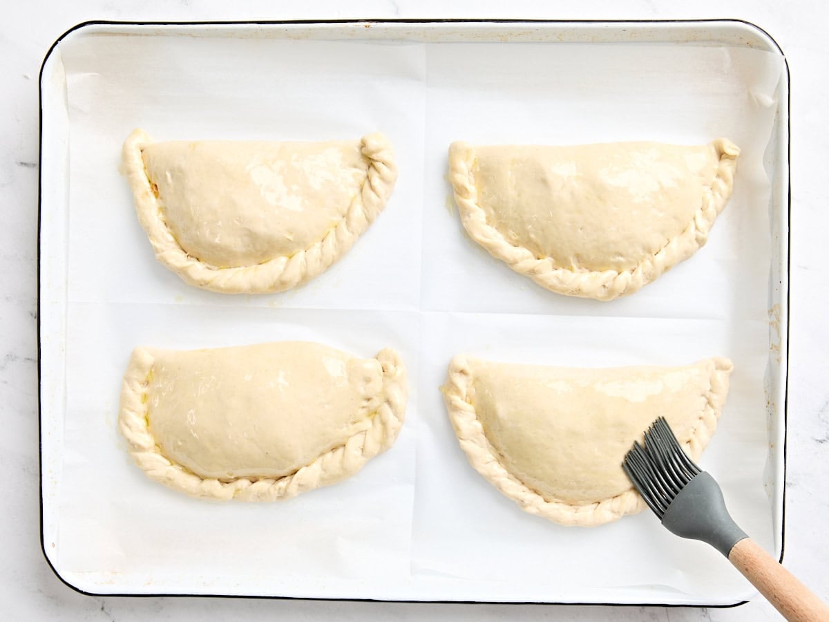 Four filled calzones on a parchment lined baking sheet being brushed with olive oil before baking.