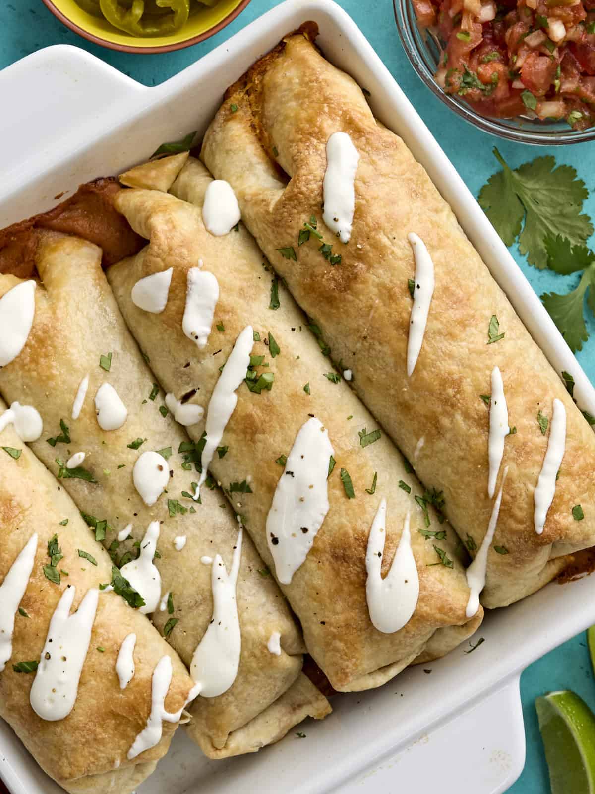 Overhead view of baked chicken chimichangas in a baking dish topped with sour cream and fresh herbs.
