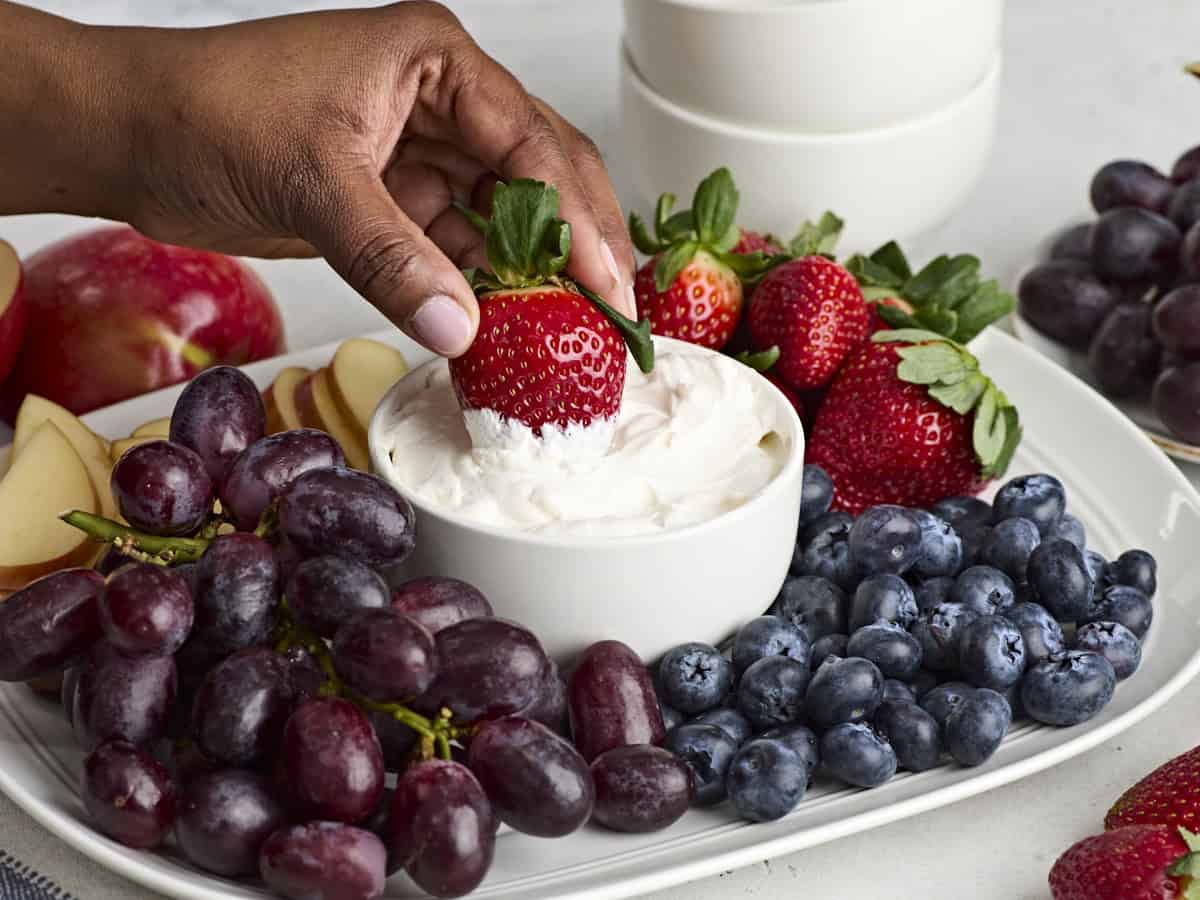 Side view of a bowl of fruit dip on a platter of fruit, with a hand dipping a strawberry in.