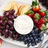 Overhead view of a bowl of homemade fruit dip surrounded by grapes, apple slices, blueberries, and strawberries.