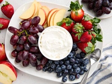 Overhead view of a bowl of homemade fruit dip surrounded by grapes, apple slices, blueberries, and strawberries.