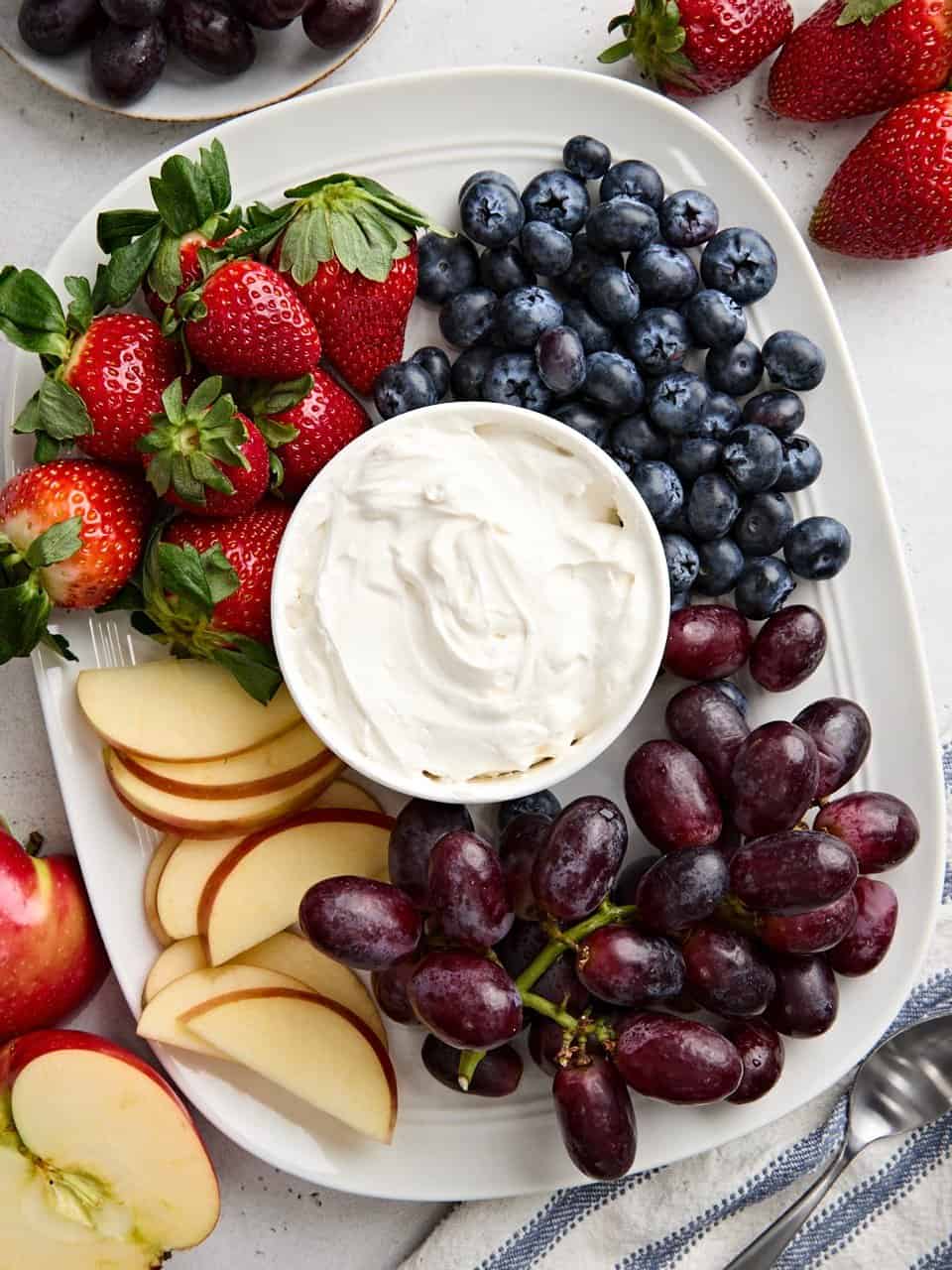 Overhead view of a bowl of homemade fruit dip surrounded by grapes, apple slices, blueberries, and strawberries.