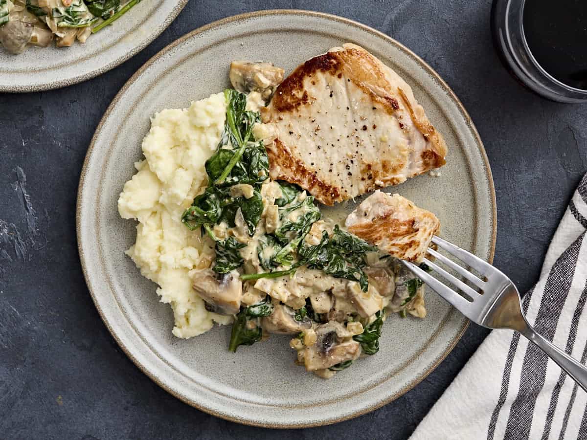 Overhead view of a creamy pork chop in a mushroom and spinach sauce with mashed potatoes, with a fork taking some.
