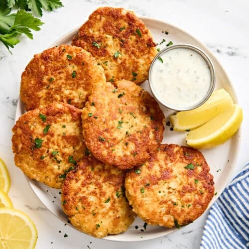 Overhead view of homemade salmon patties on a plate with tartar sauce and lemon wedges.