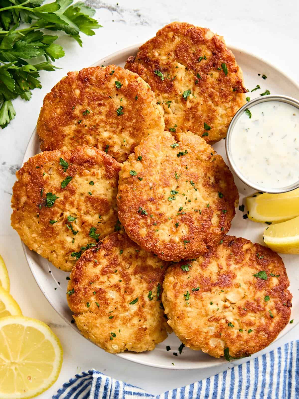 Overhead view of salmon patties on a plate with tartar sauce and lemon wedges.