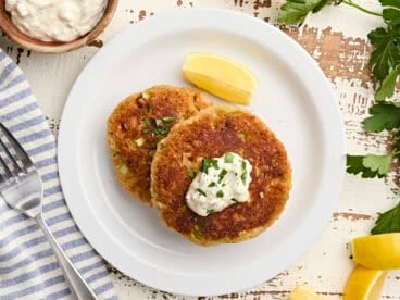 Overhead view of two homemade crab cakes on a plate with a lemon wedge and tartar sauce.