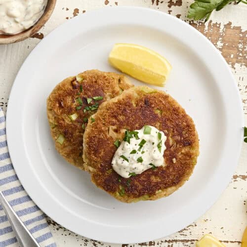 Overhead view of two homemade crab cakes on a plate with a lemon wedge and tartar sauce.