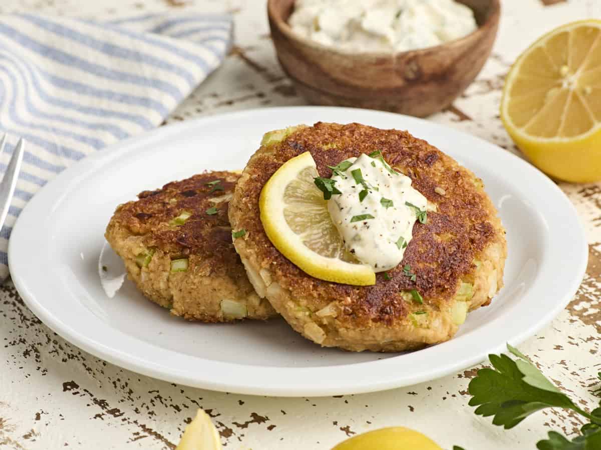 Side view of two canned crab cakes on a plate with a lemon wedge and tartar sauce.