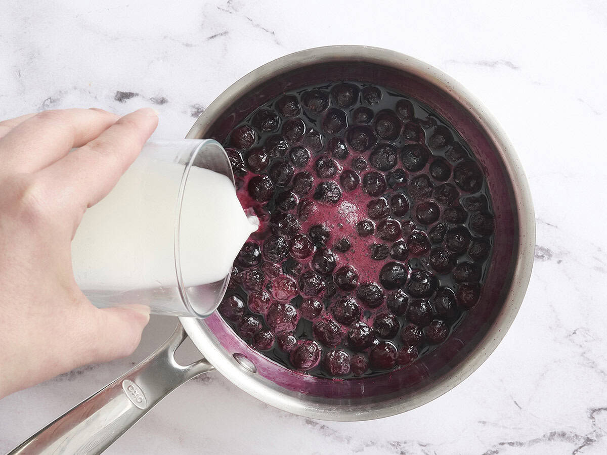 A cornstarch slurry being poured into blueberry sauce in a sauce pot.