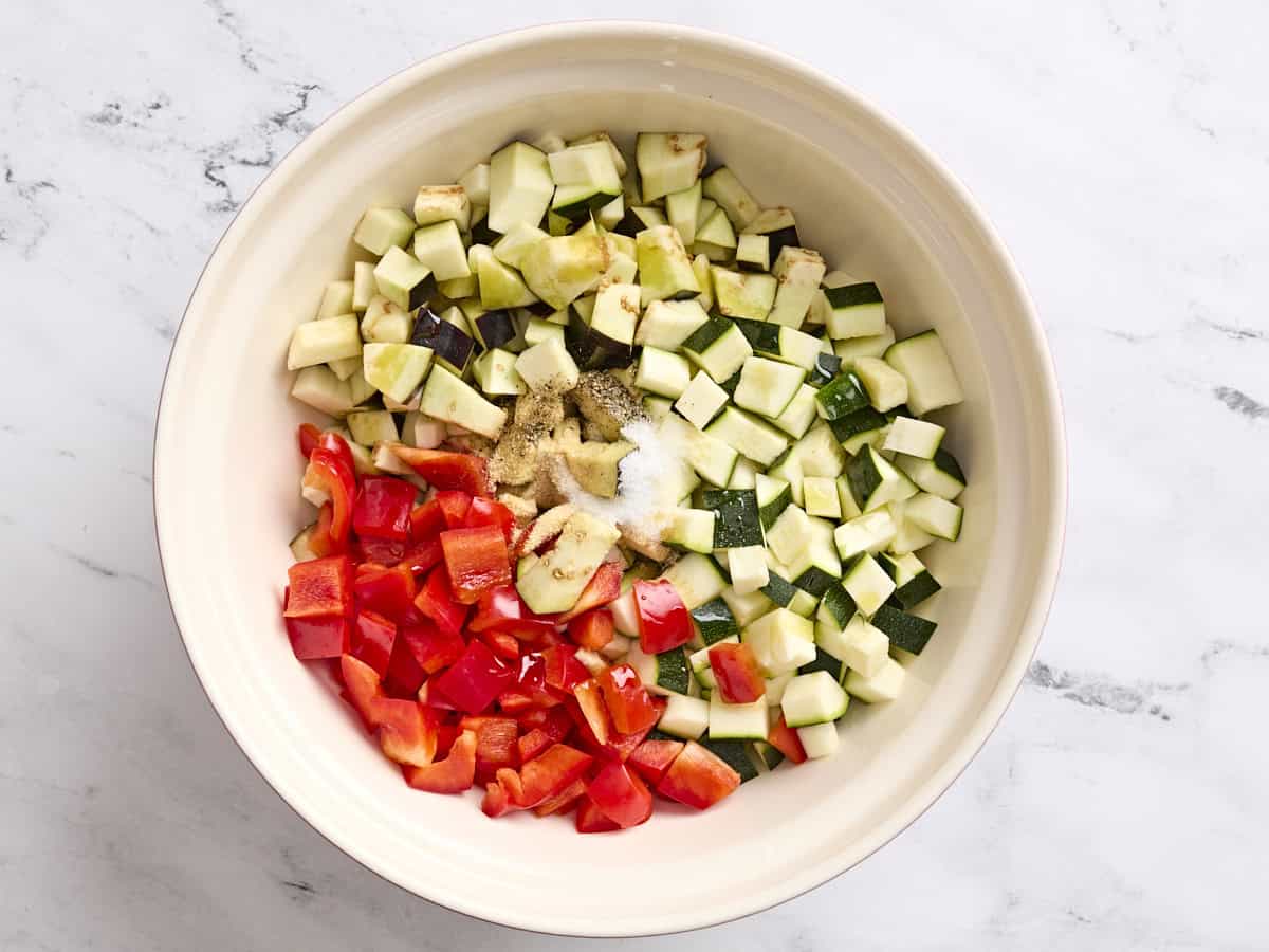 Chopped red bell pepper, zucchini and eggplant in a mixing bowl with seasonings.