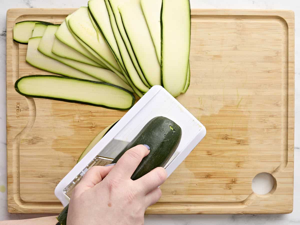 A hand slicing a zucchini with a mandolin slicer.