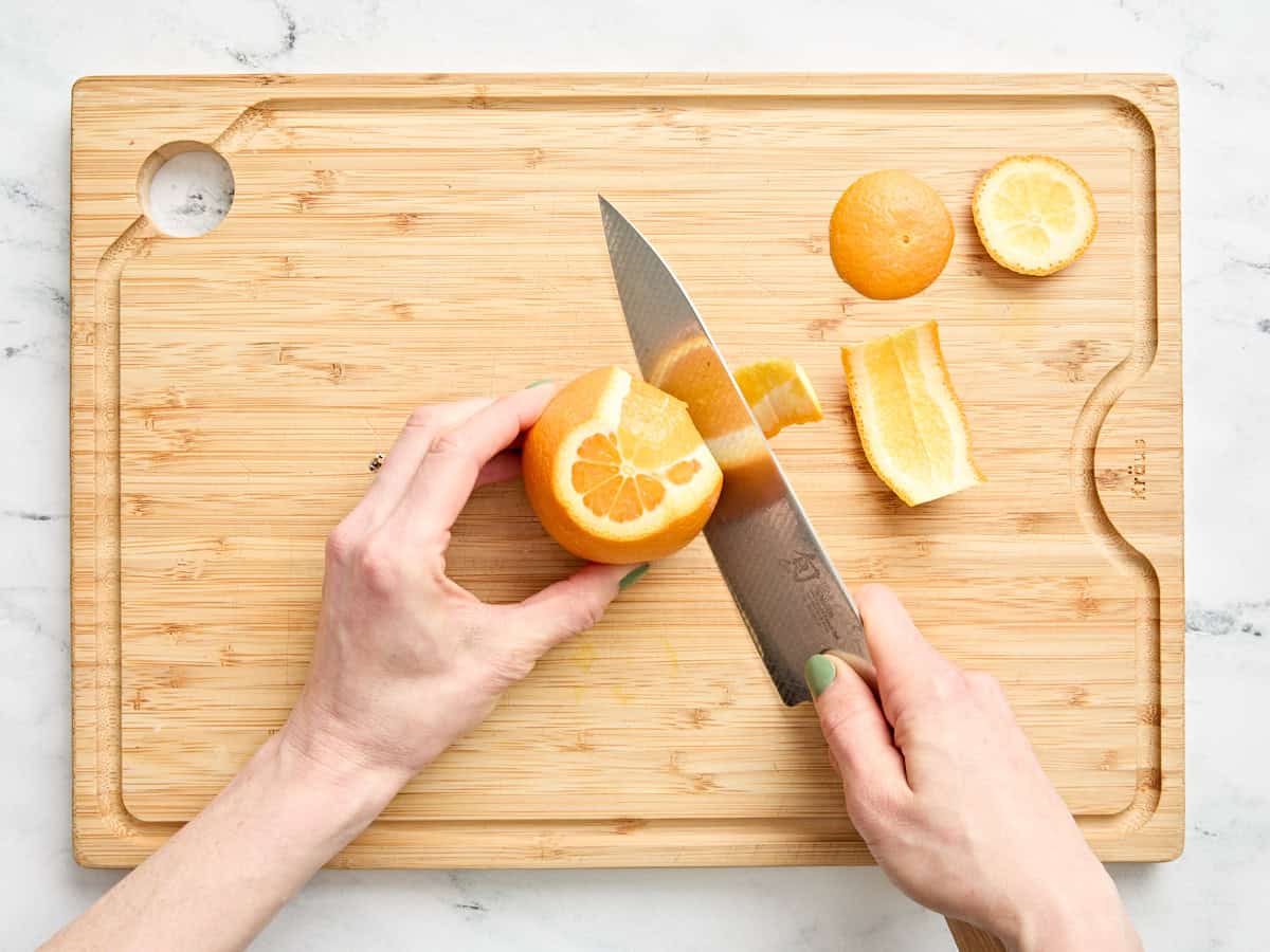 A knife cutting the skin off an orange on a wooden cutting board.