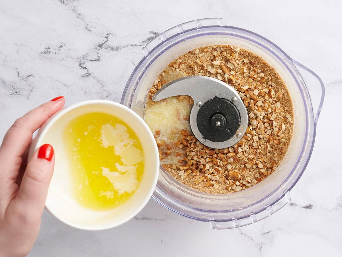 Melted butter being poured into a food processor with crushed pretzels and brown sugar.