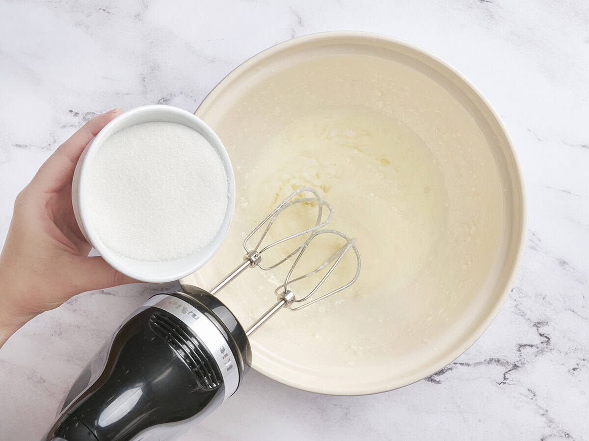 Granulated sugar being poured into a bowl of whipped butter and oil.
