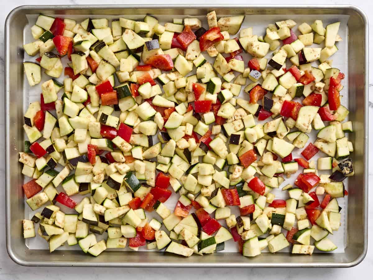 Chopped zucchini, eggplant and red bell pepper on a parchment lined baking sheet.