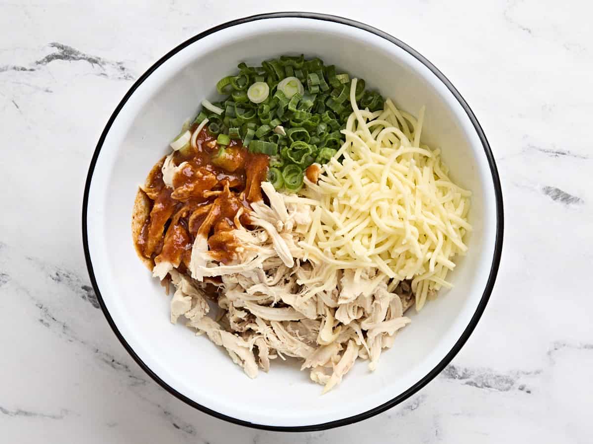 Green onions, shredded cheese, shredded chicken, and enchilada sauce in a bowl.