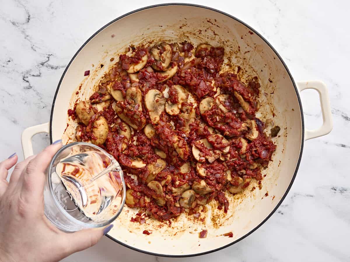 A person holding a glass of water over a pan of mushrooms and tomato paste.
