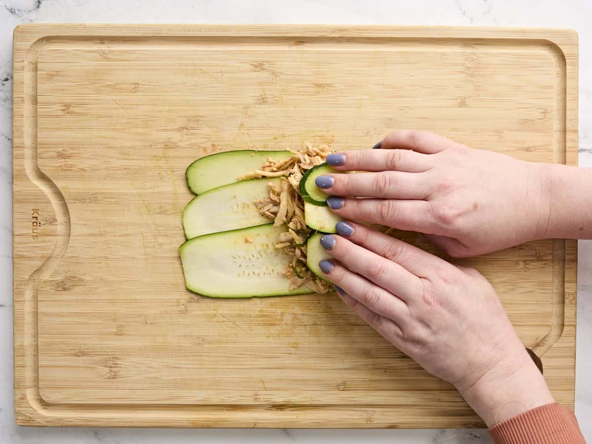Hands rolling a zucchini enchilada on a wooden cutting board.