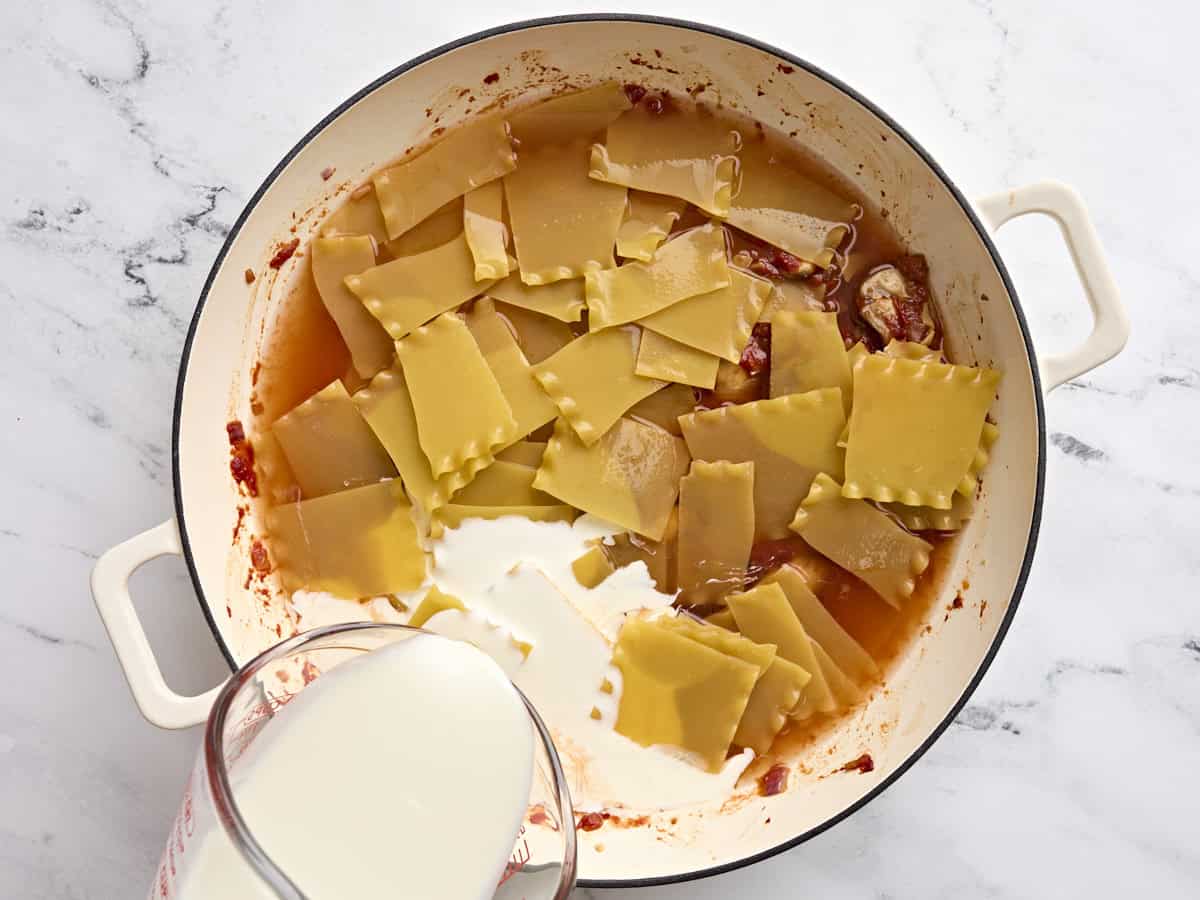 A pan filled with broken lasagna sheets and creamy being poured into it.