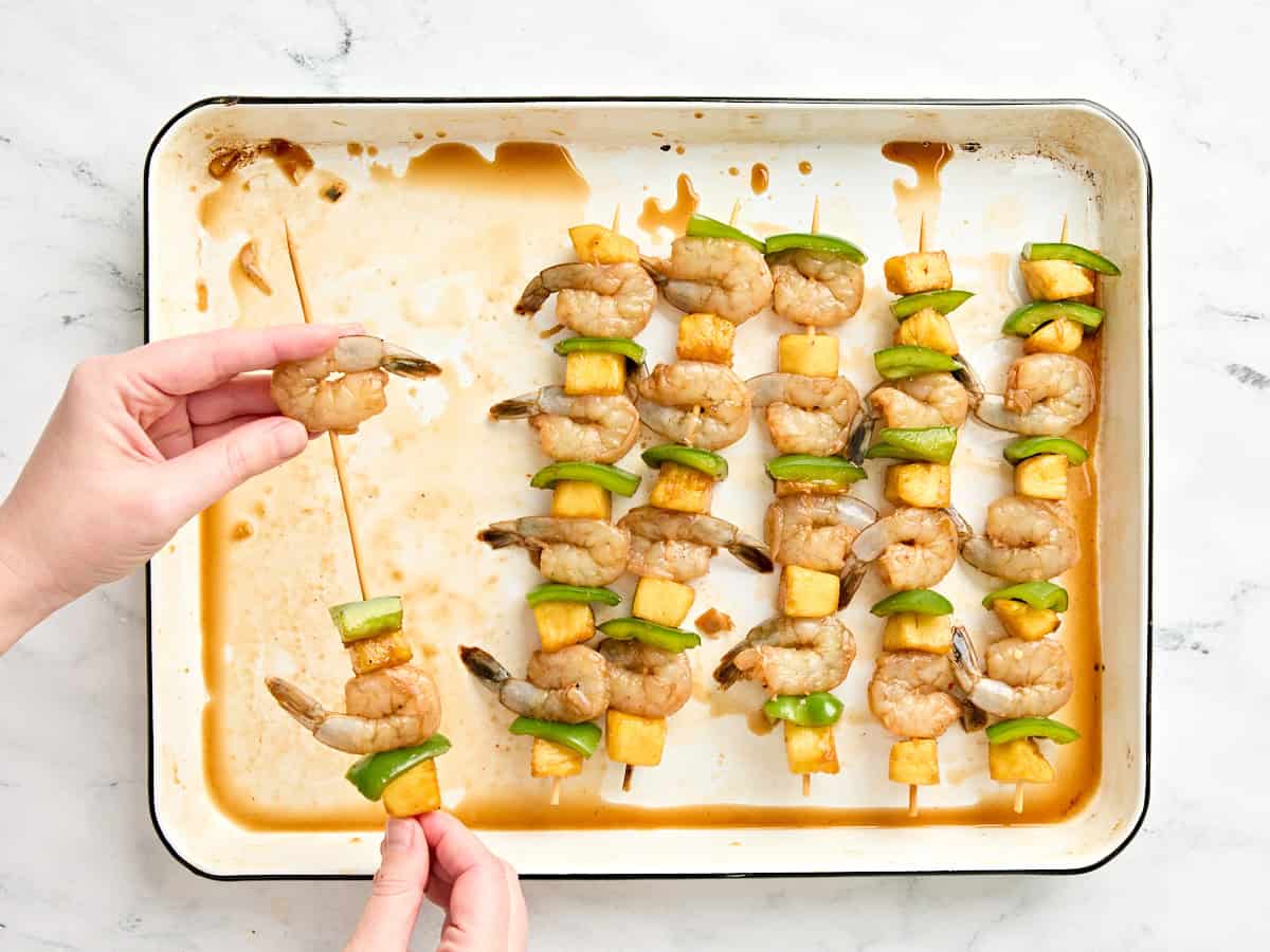 Hands threading raw shrimp, pineapple chunks, and bell pepper onto wooden skewers.