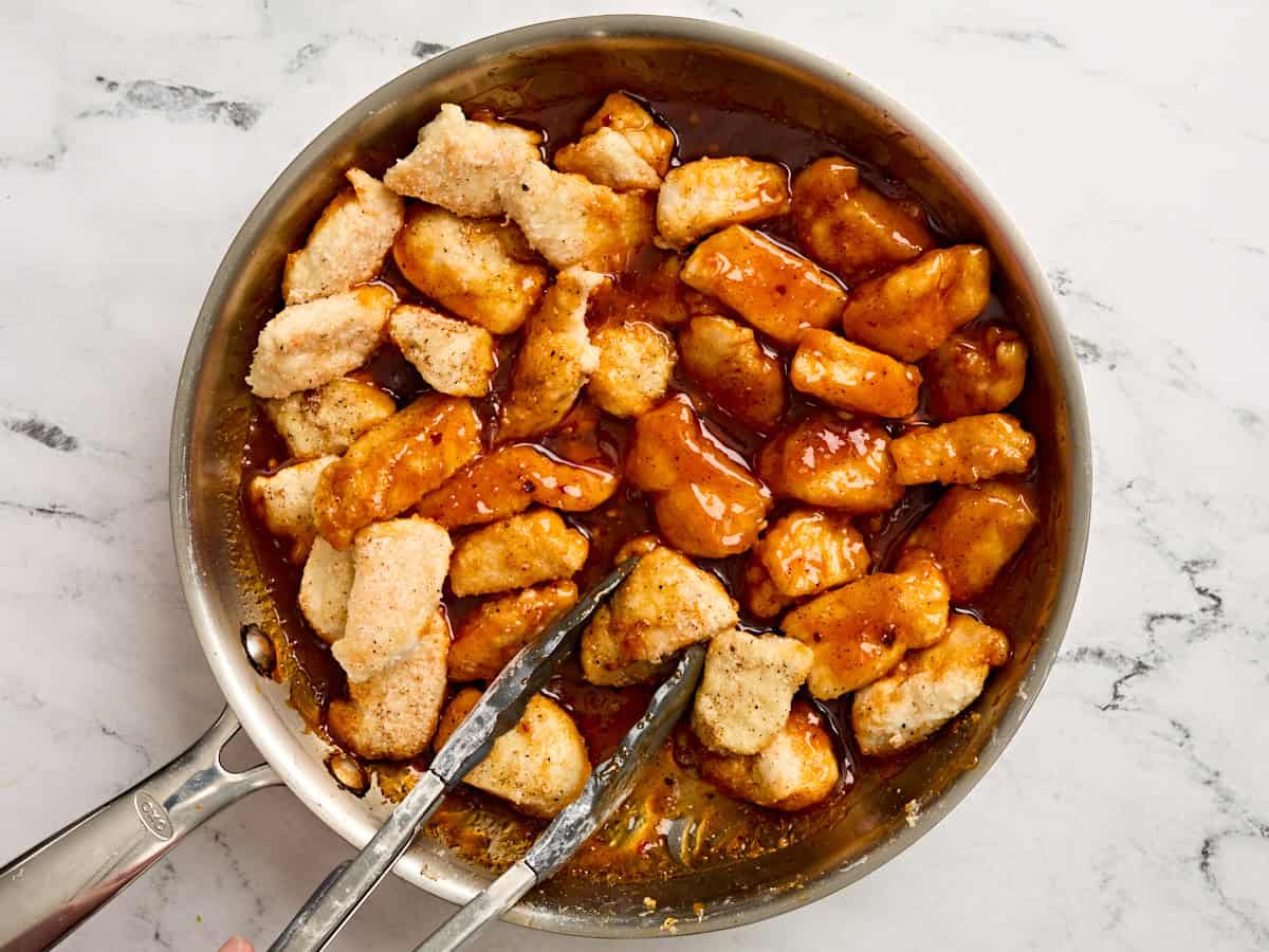 Kitchen tongs flipping over fire cracker chicken in a skillet.