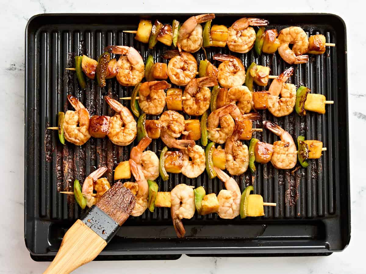 A brush basting pineapple and shrimp skewers with marinade on a grill.
