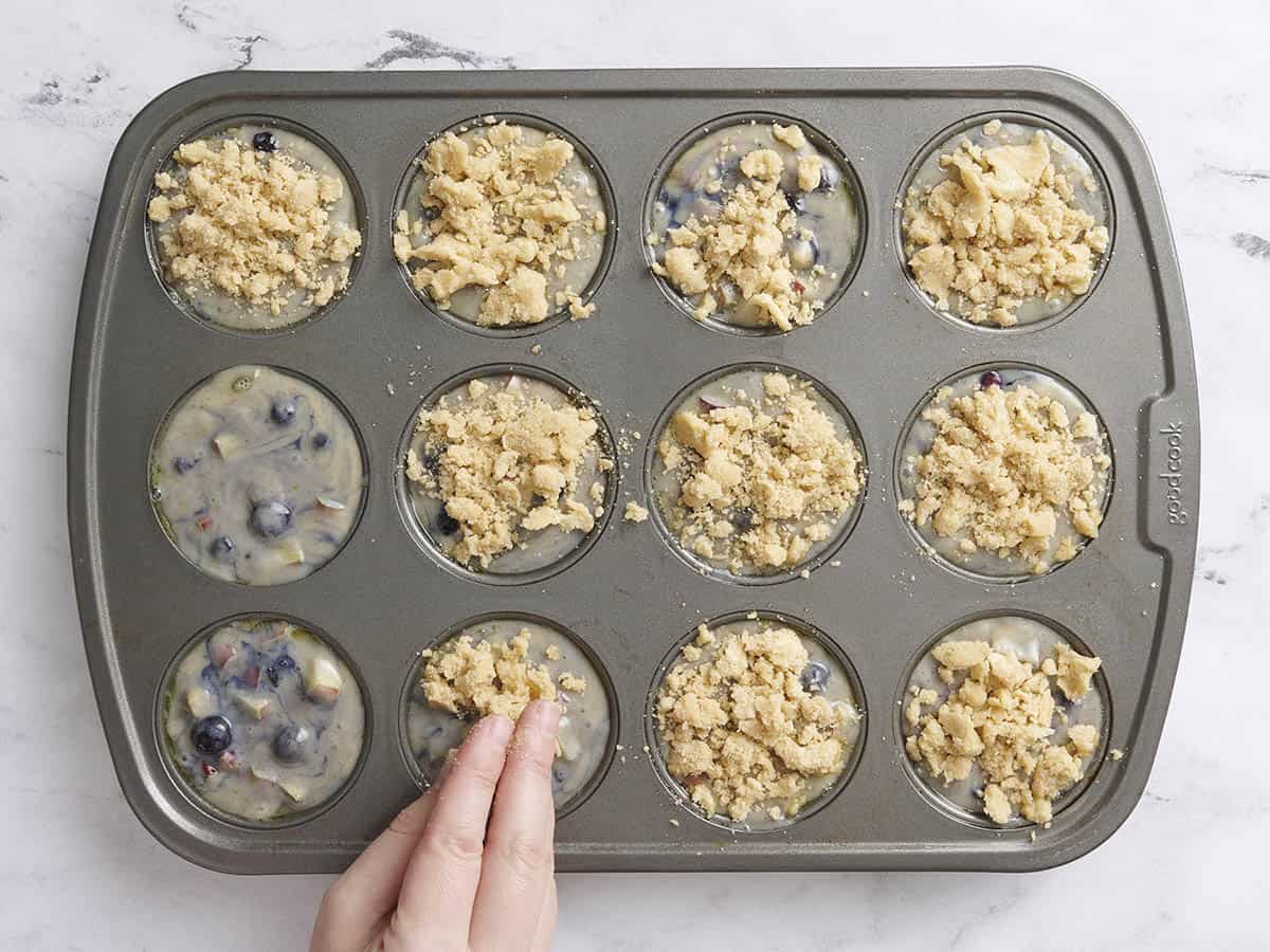 A hand topping blueberry rhubarb muffin batter with streusel topping in a mixing bowl.
