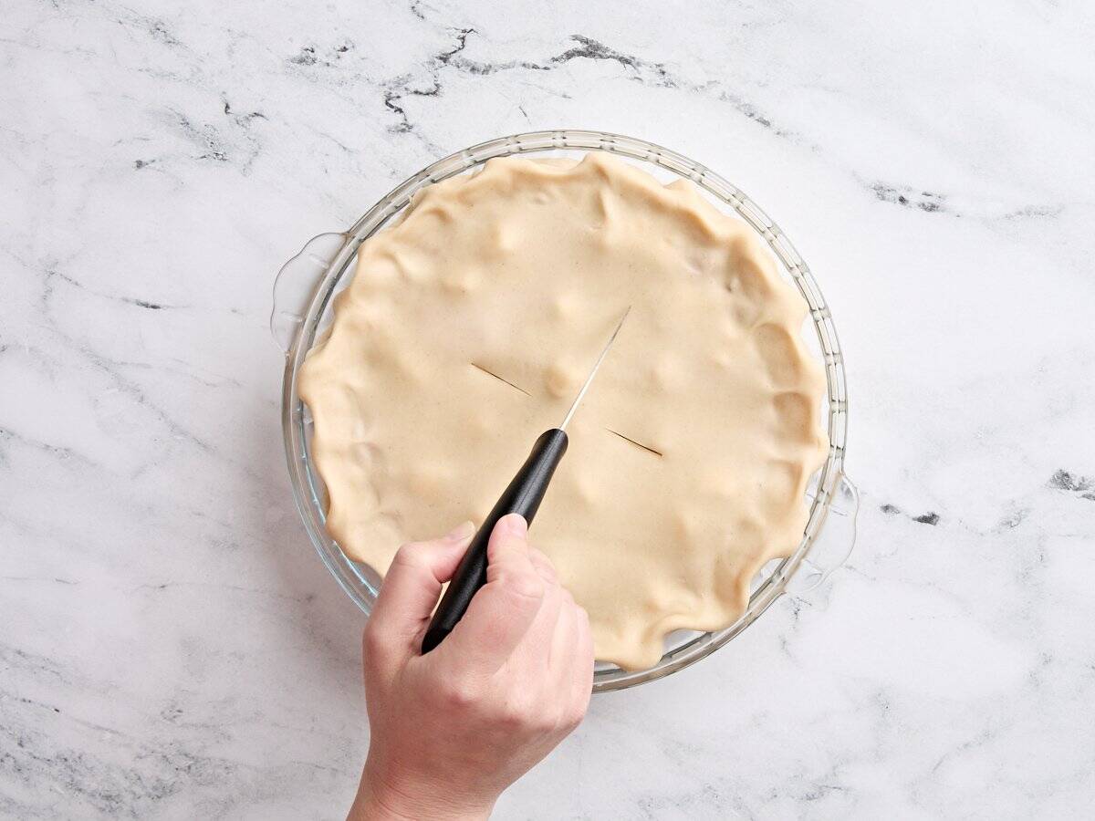 A knife slicing a slit in an unbaked rhubarb strawberry pie.