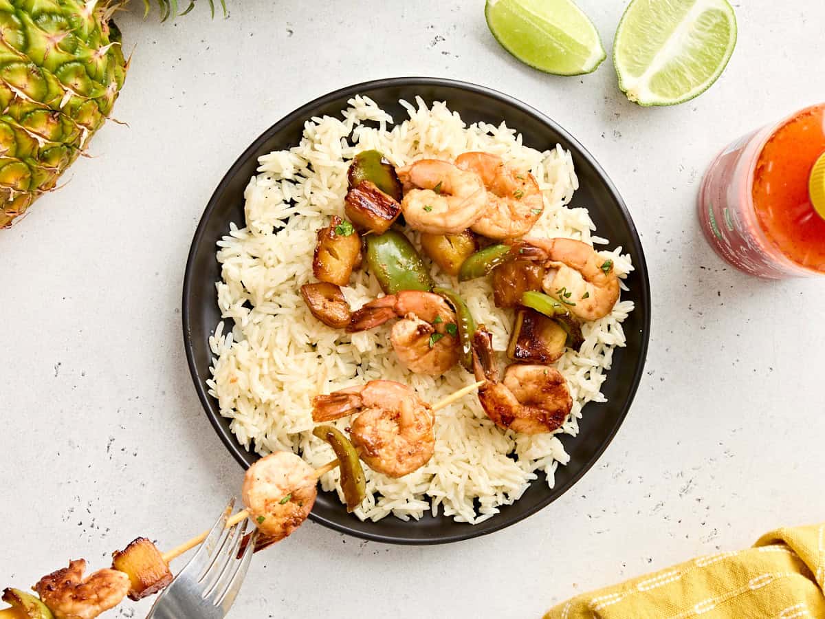 Grilled pineapple and shrimp being pulled off a wooden skewer onto a plate of rice.