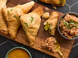 Overhead view of homemade puff pastry samosas on a wooden board with a bowl of onion chutney.