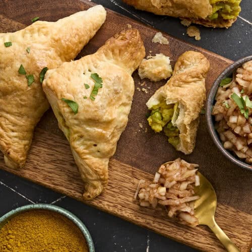 Overhead view of homemade puff pastry samosas on a wooden board with a bowl of onion chutney.
