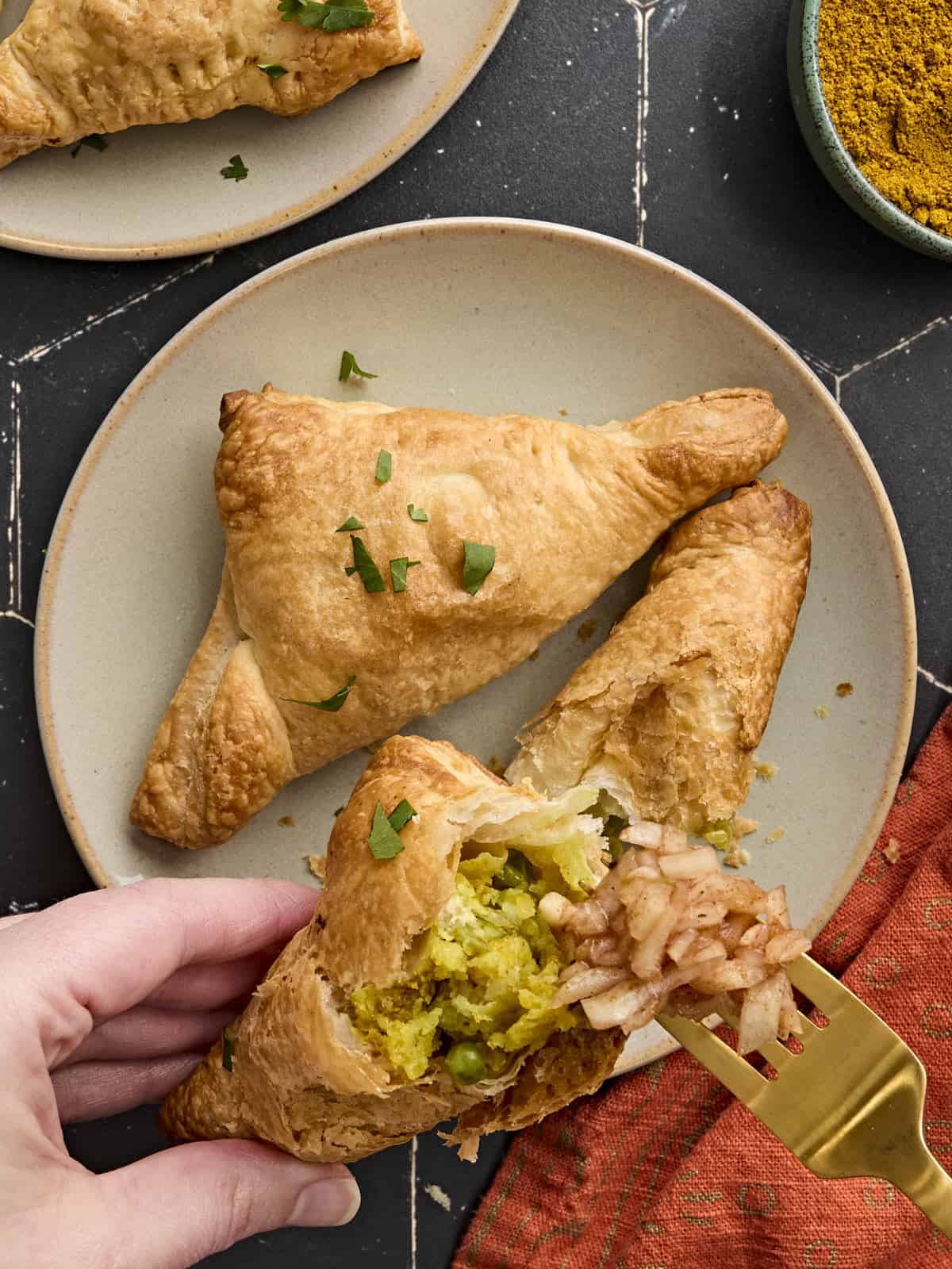 Hands holding half a puff pastry samosa with a fork topping it with onion chutney.