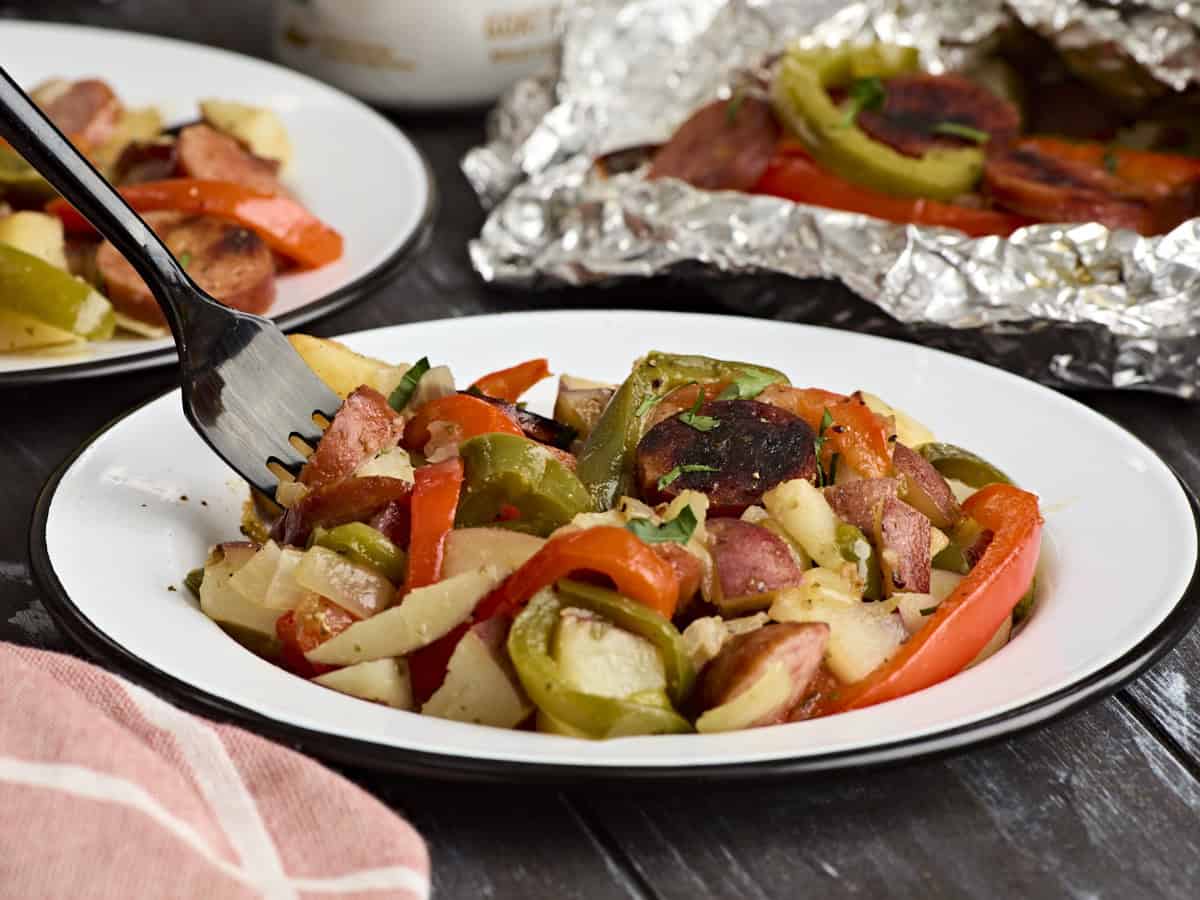 Side view of a plate of grilled sausage and peppers with grilled sausage and pepper foil packets in the background.